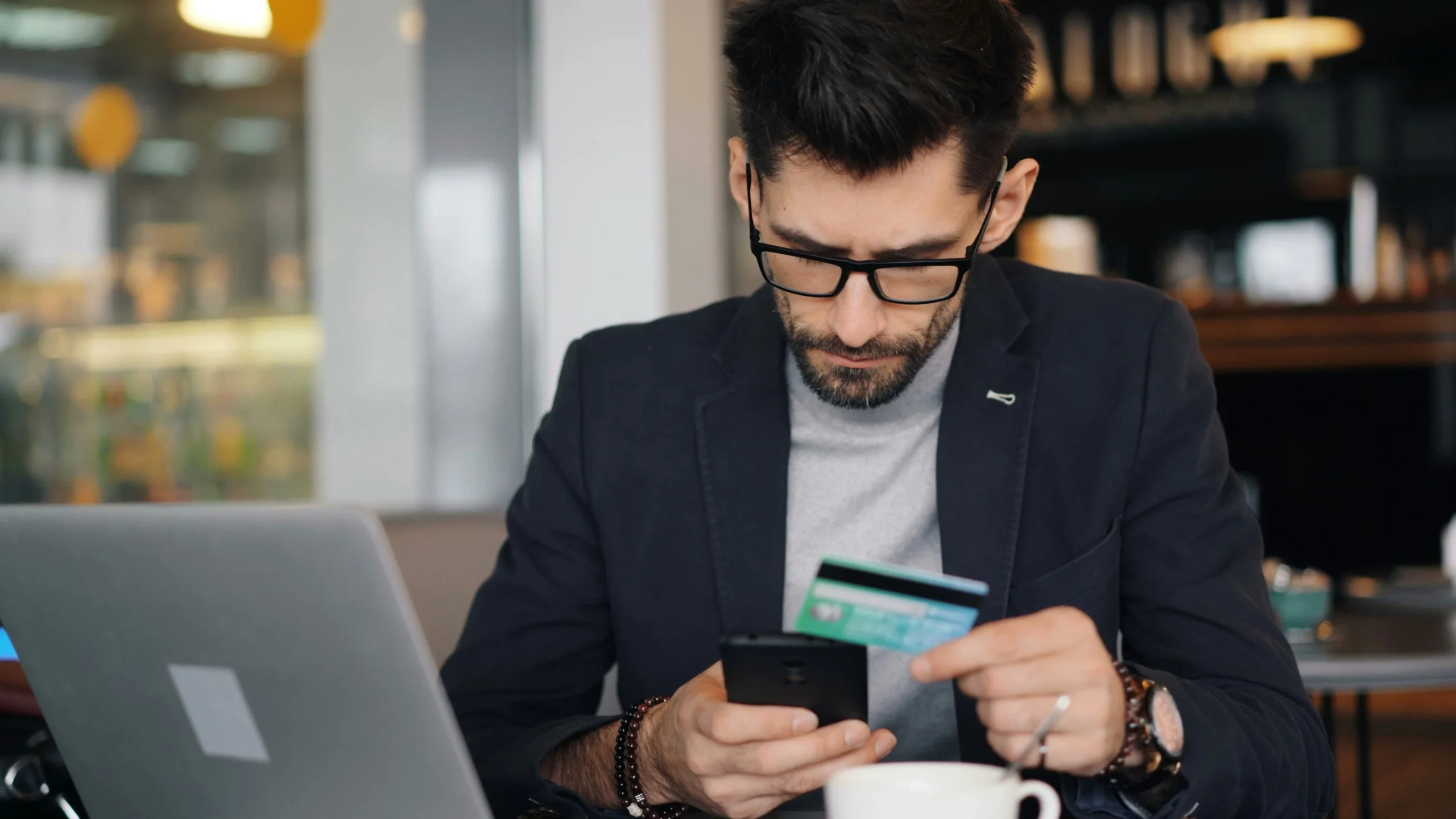 Hombre joven con gafas y chaqueta revisa su teléfono móvil mientras sostiene una tarjeta bancaria, sentado frente a un portátil y una taza de café en un entorno de cafetería.