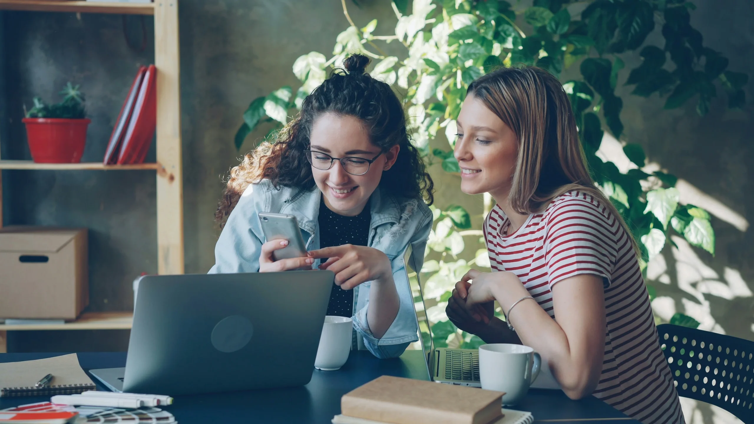 Dos mujeres jóvenes sonríen mientras miran juntas la pantalla de un teléfono móvil en un entorno de oficina informal. Frente a ellas hay un ordenador portátil, tazas de café y cuadernos, transmitiendo un ambiente de trabajo colaborativo y relajado.