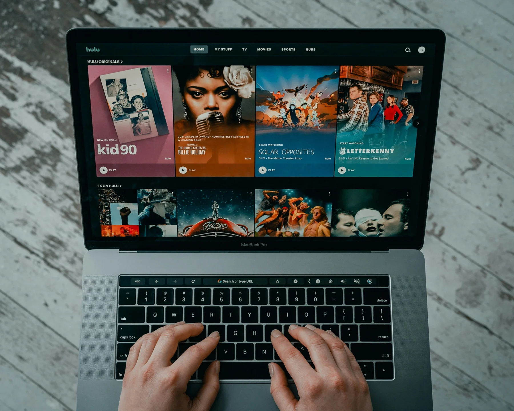 Persona usando un ordenador portátil MacBook Pro en un ambiente interior, navegando el sitio web de Hulu, donde se muestran varias recomendaciones de series y películas, como “kid 90”, “Solar Opposites” y “Letterkenny”. Las manos de la persona están en el teclado, listas para escribir o seleccionar contenido.