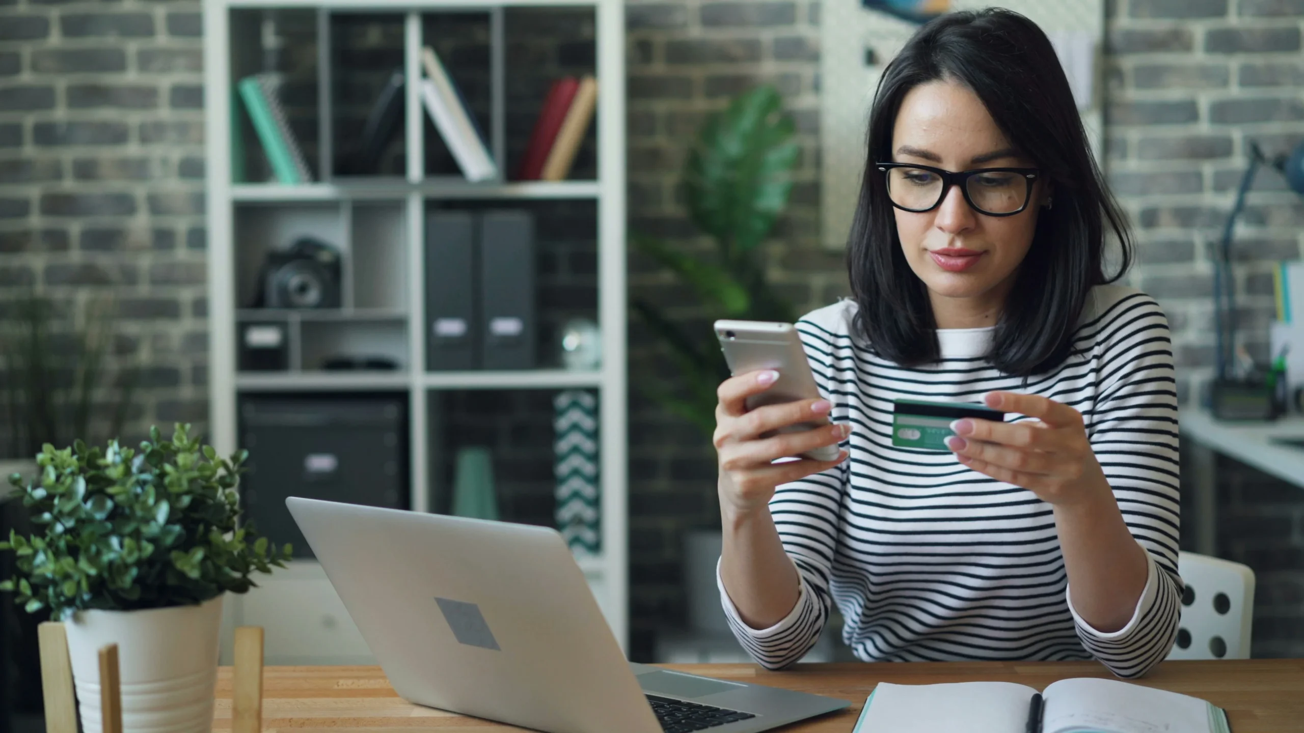 Mujer ingresando datos de su tarjeta en un teléfono móvil mientras utiliza un ordenador, ilustrando el uso de pagos digitales seguros.