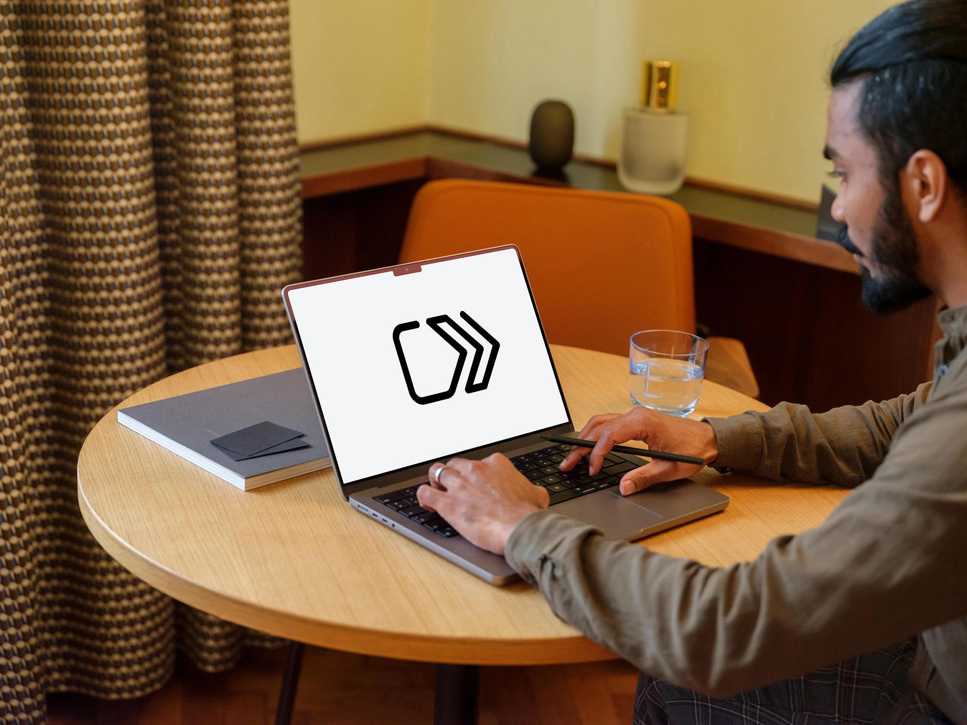 Persona sentada en una mesa redonda de madera, utilizando un ordenador portátil que muestra el logotipo de Click to Pay en la pantalla. Sobre la mesa hay una libreta cerrada con una tarjeta encima y un vaso de agua. Al fondo se observa una silla, cortinas y algunos objetos decorativos.
