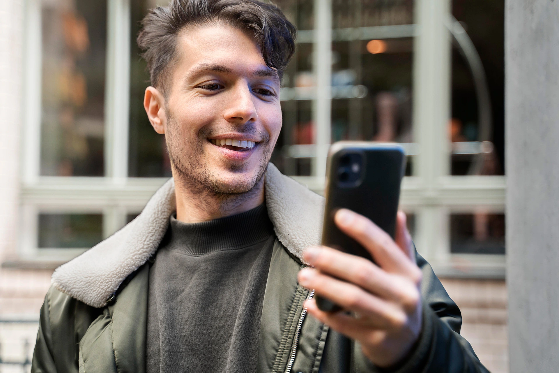 Primer plano de un hombre joven con un teléfono móvil en la mano. Lleva cazadora verde con cuello de borreguillo y camiseta negra. El fondo muestra una ventana amplia y ambiente urbano.