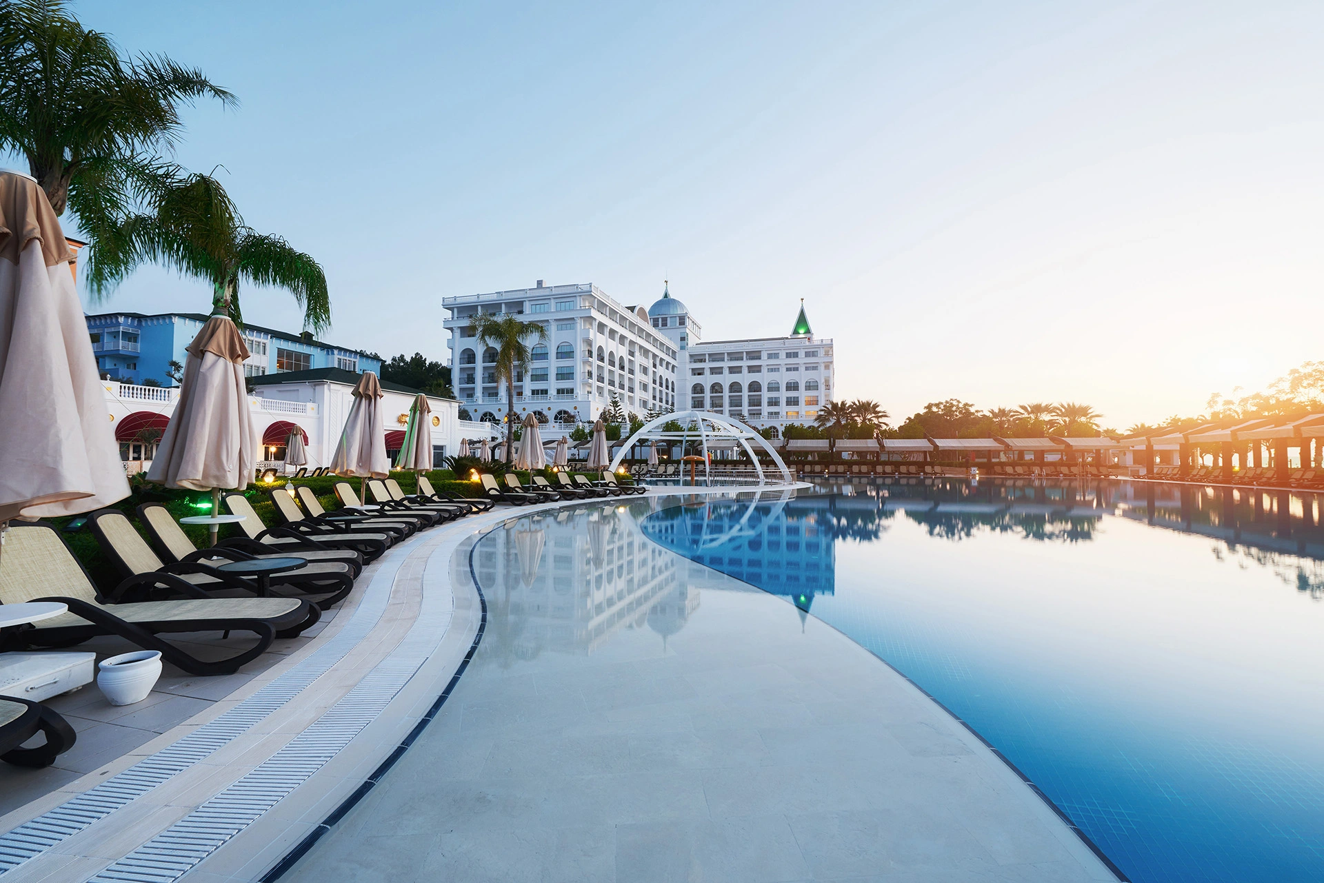 Piscina de hotel grande y lujosa rodeada de tumbonas y sombrillas beige, con palmeras decorativas a los lados. Al fondo se observa un edificio blanco de varios pisos con arquitectura elegante y cúpulas, todo bajo una luz suave de atardecer o amanecer.